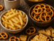 Verschiedene salzige Snacks wie Pommes, Salzstangen, Brezeln, Cracker und Chips in Schälchen auf einem Holztisch arrangiert